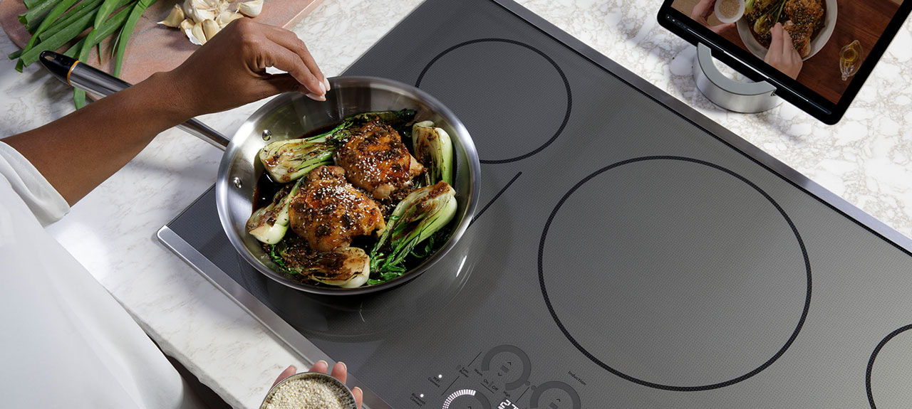 Person cooking on an induction cooktop with a bowl of food and a screen nearby showing Hestan Cue Guided Cooking.