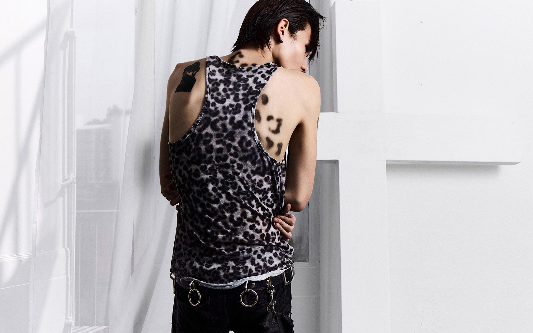 Model with short hair wearing a leopard-print tank top posing near a window in a soft-lit studio