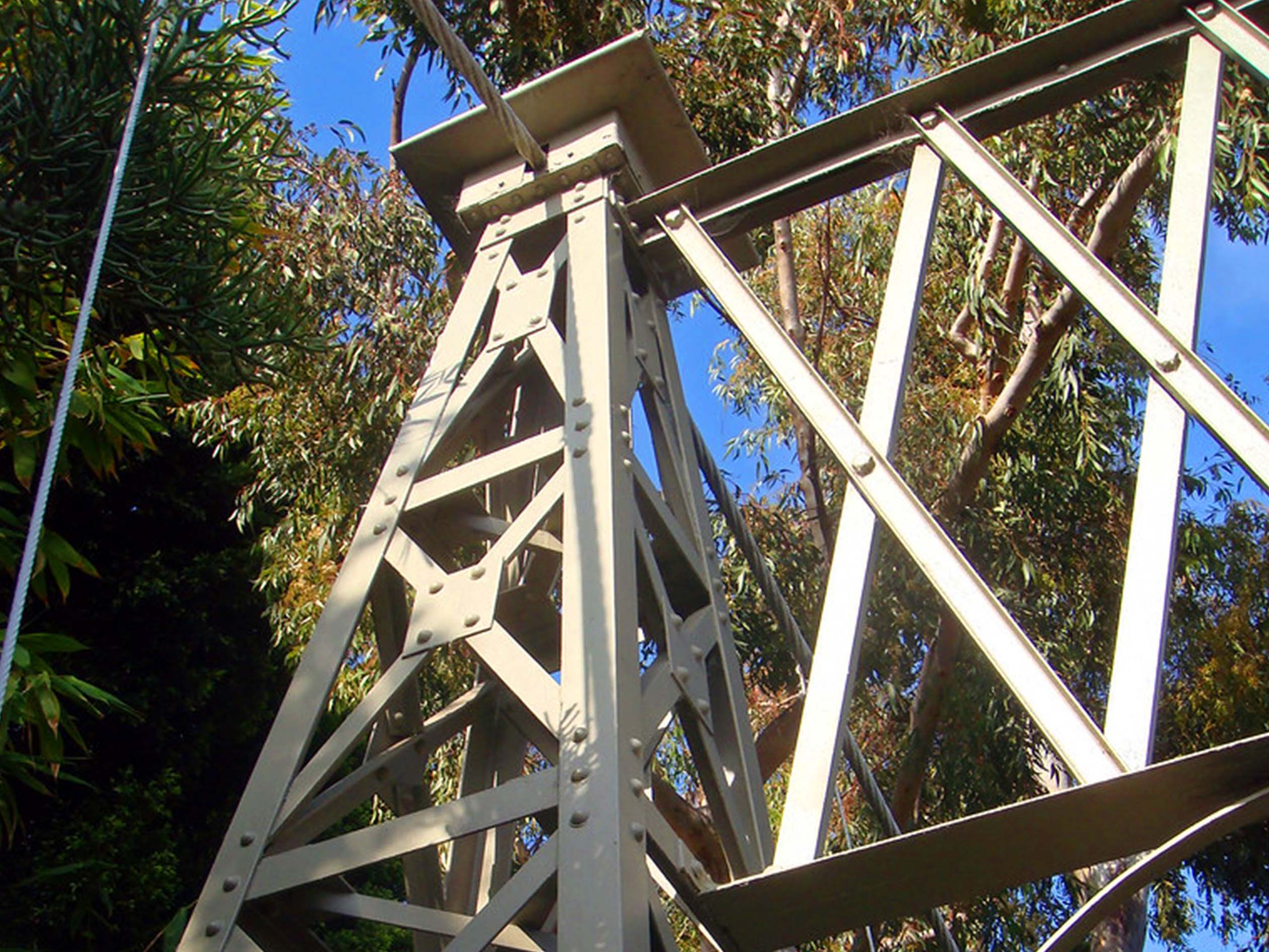 A closeup photo of metal beams holding up the suspension lines on spruce street suspension bridge with trees in the background.