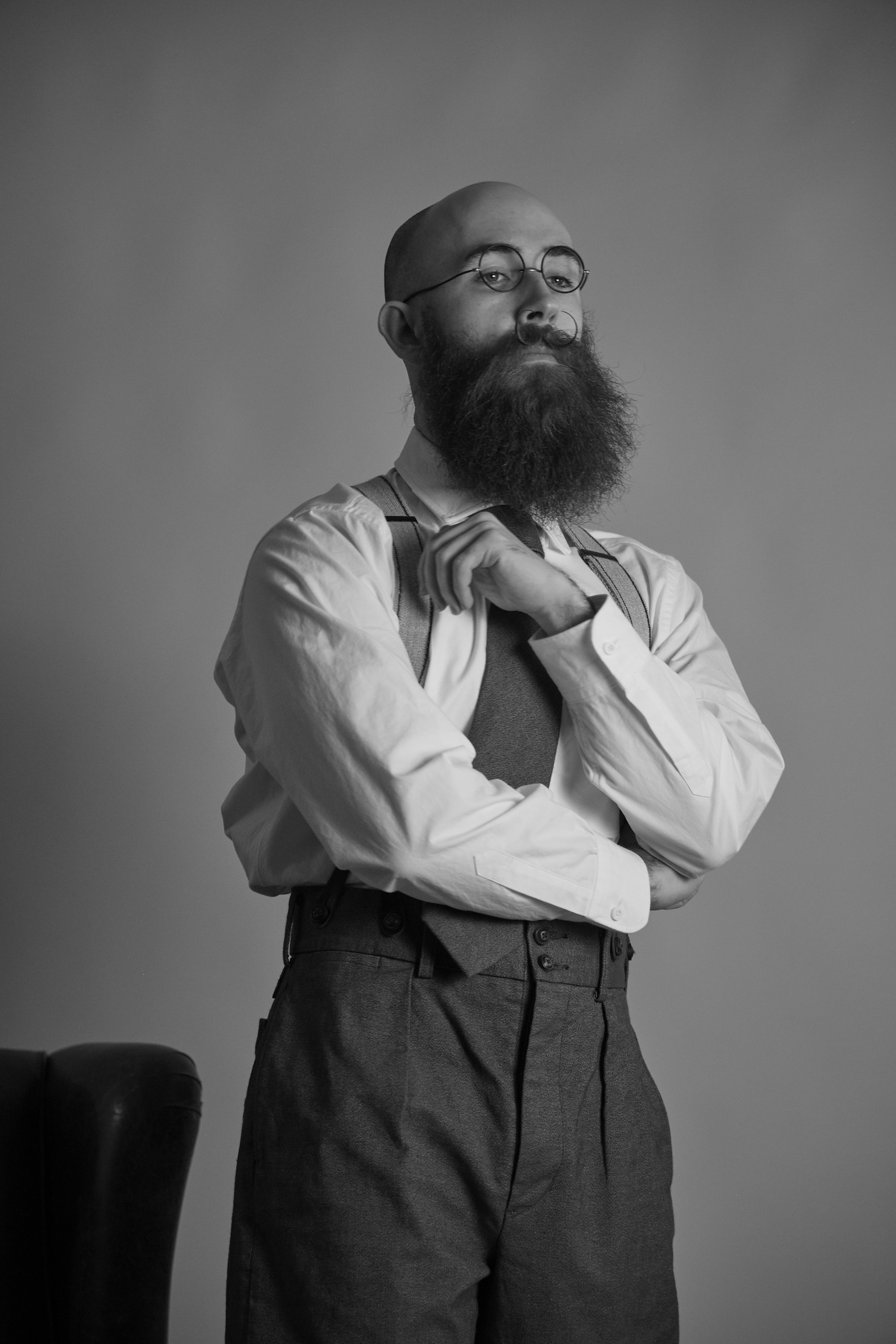 Man with a beard sitting on a chair, wearing a vest and trousers.