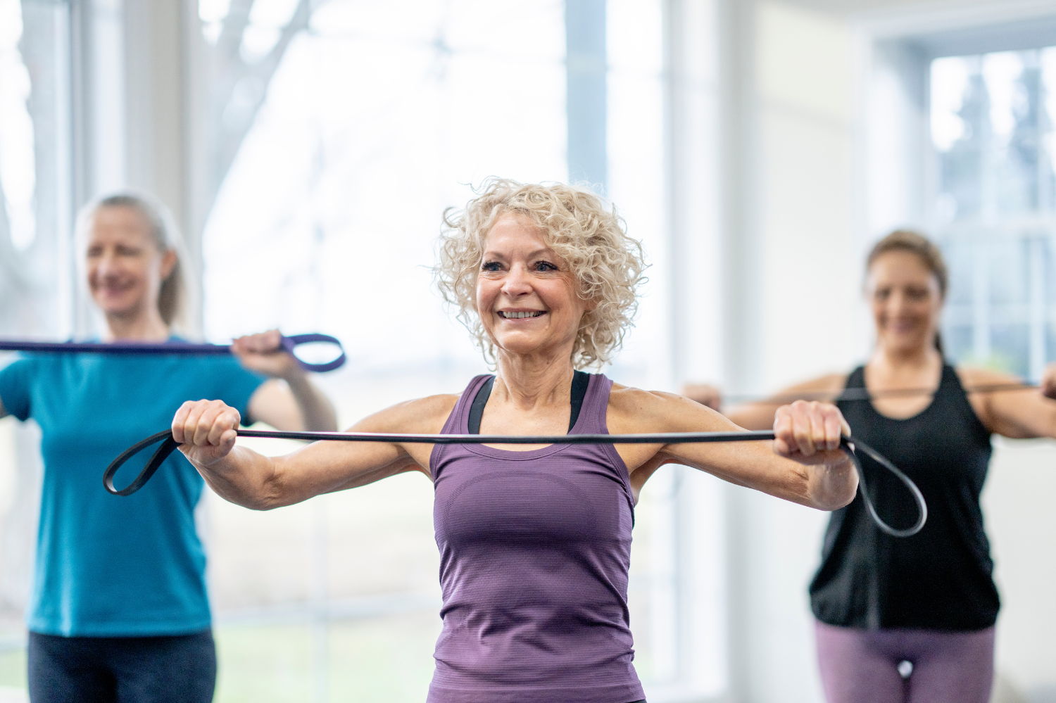 Three women exercising with resistance bands in a bright fitness studio.