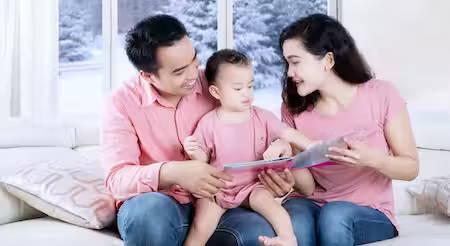 Photo of family with child sitting inside home reading a book in front of winter window