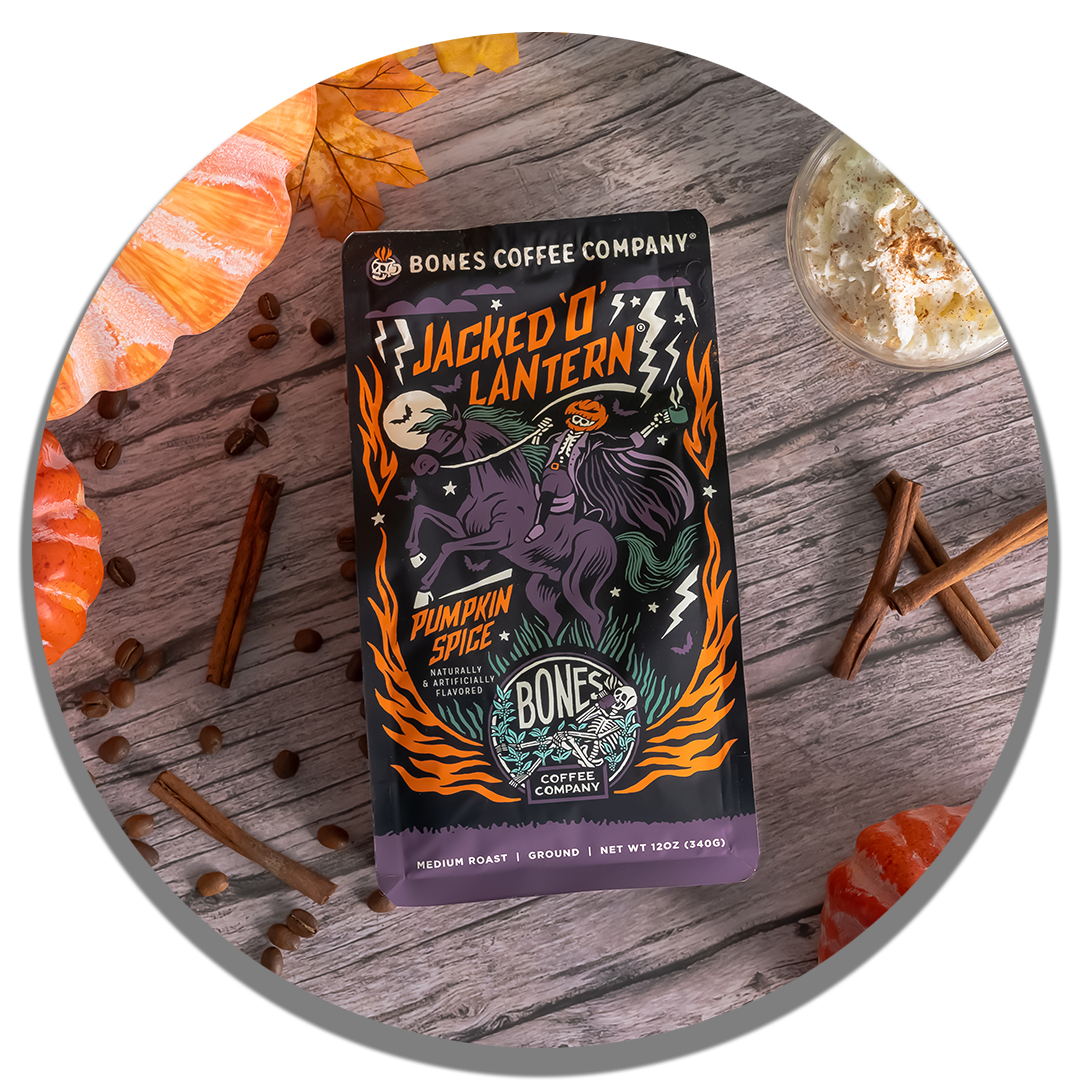 The front of a 12 ounce bag of Bones Coffee Company Jacked O’ Lantern coffee. Its flavor is pumpkin spice, and it has a skeleton with a pumpkin on its head riding a horse on the art. It is on a table with pumpkins, coffee beans, and cinnamon.
