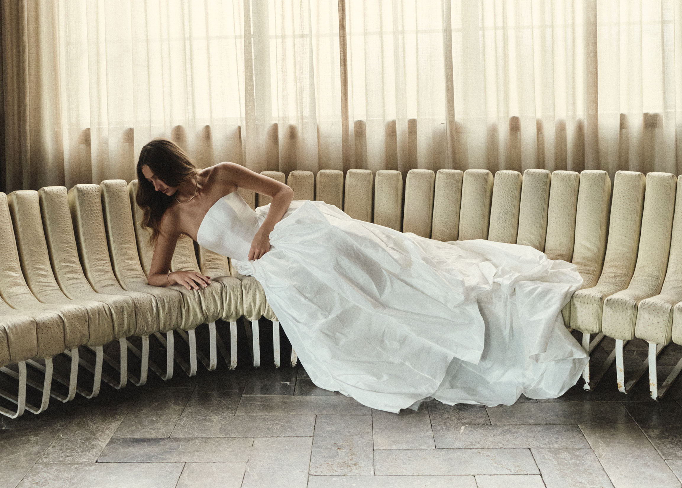 Bride in white dress lounging on curved seating in a bright room.