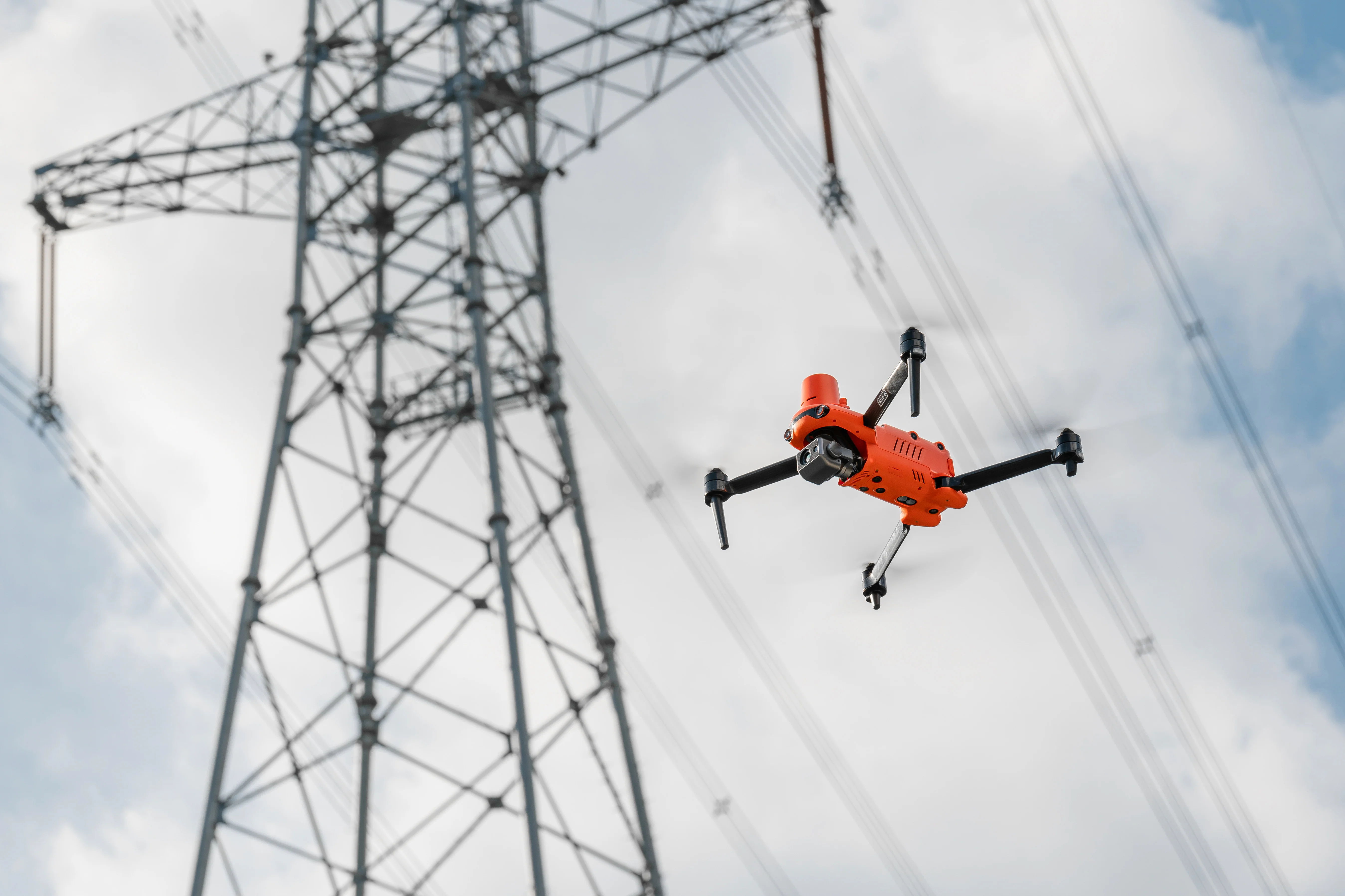 A drone flying near a power transmission tower against a cloudy sky.