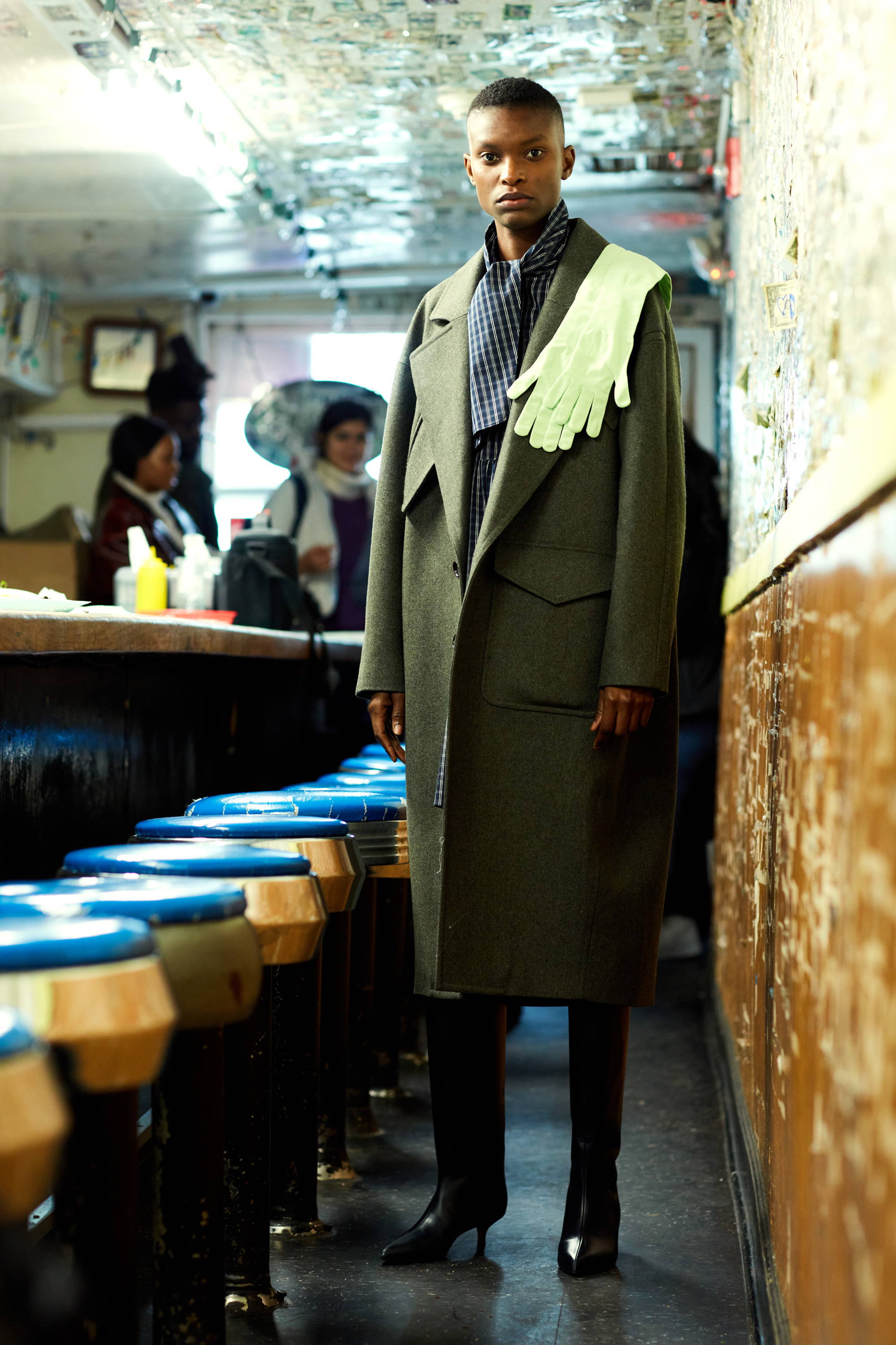 Person in a stylish coat stands in a bar with blue stools.