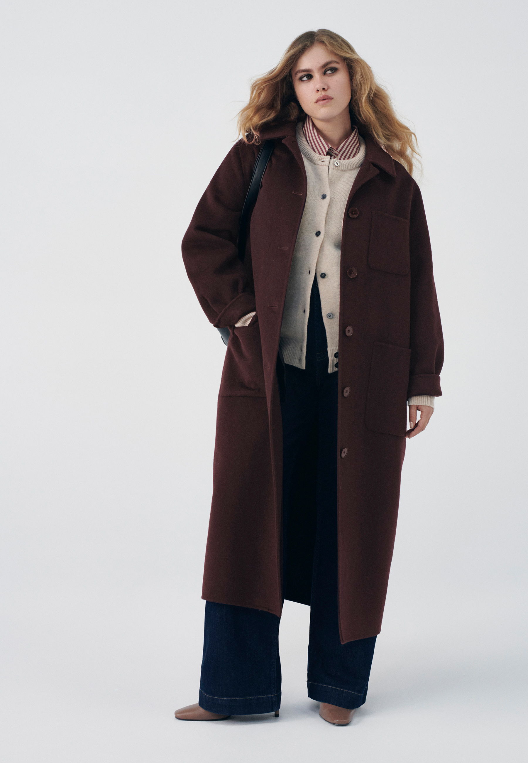 Model wearing a long brown coat over a beige sweater and dark pants.