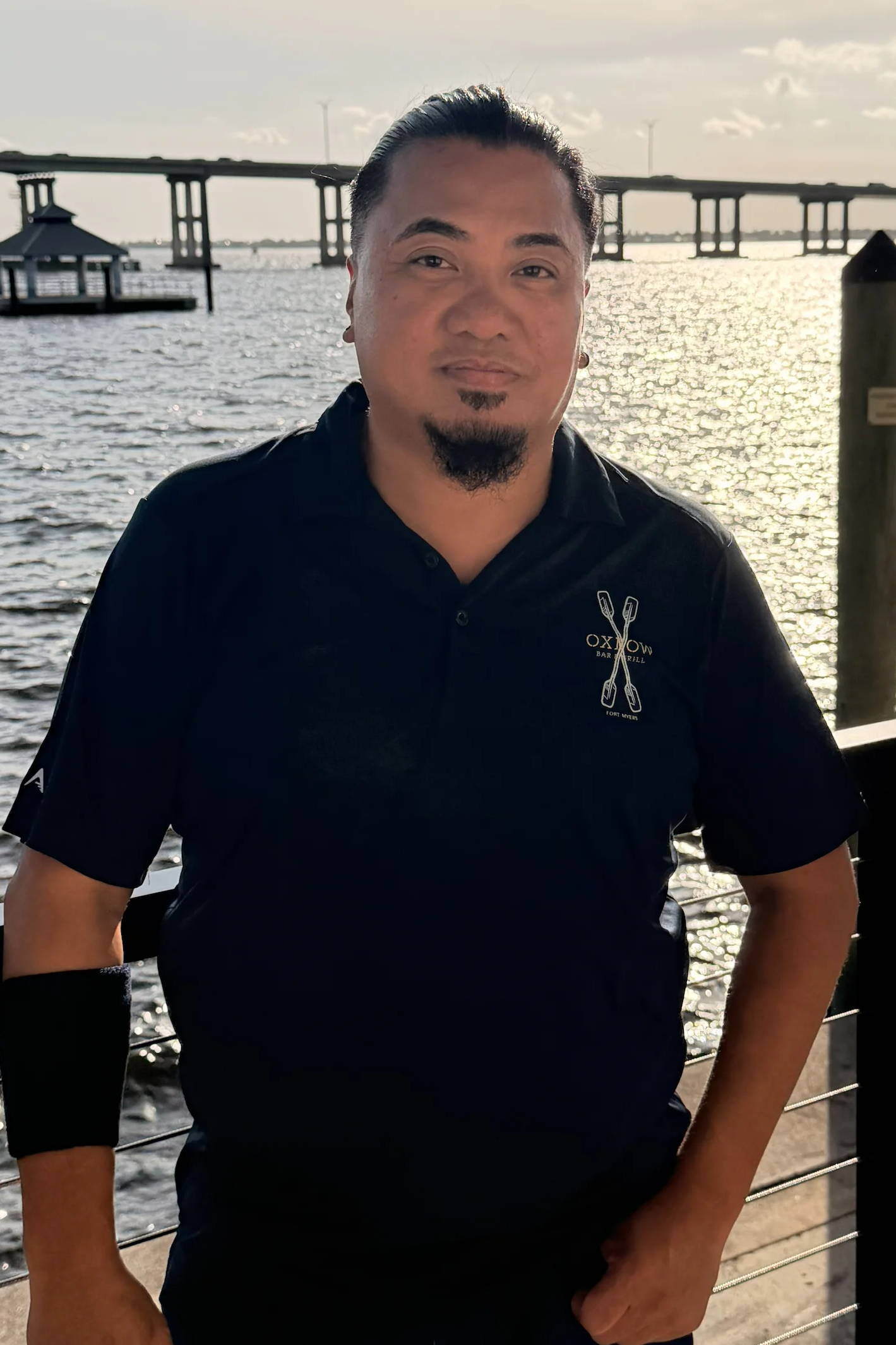A man standing by the water with a pier in the background.