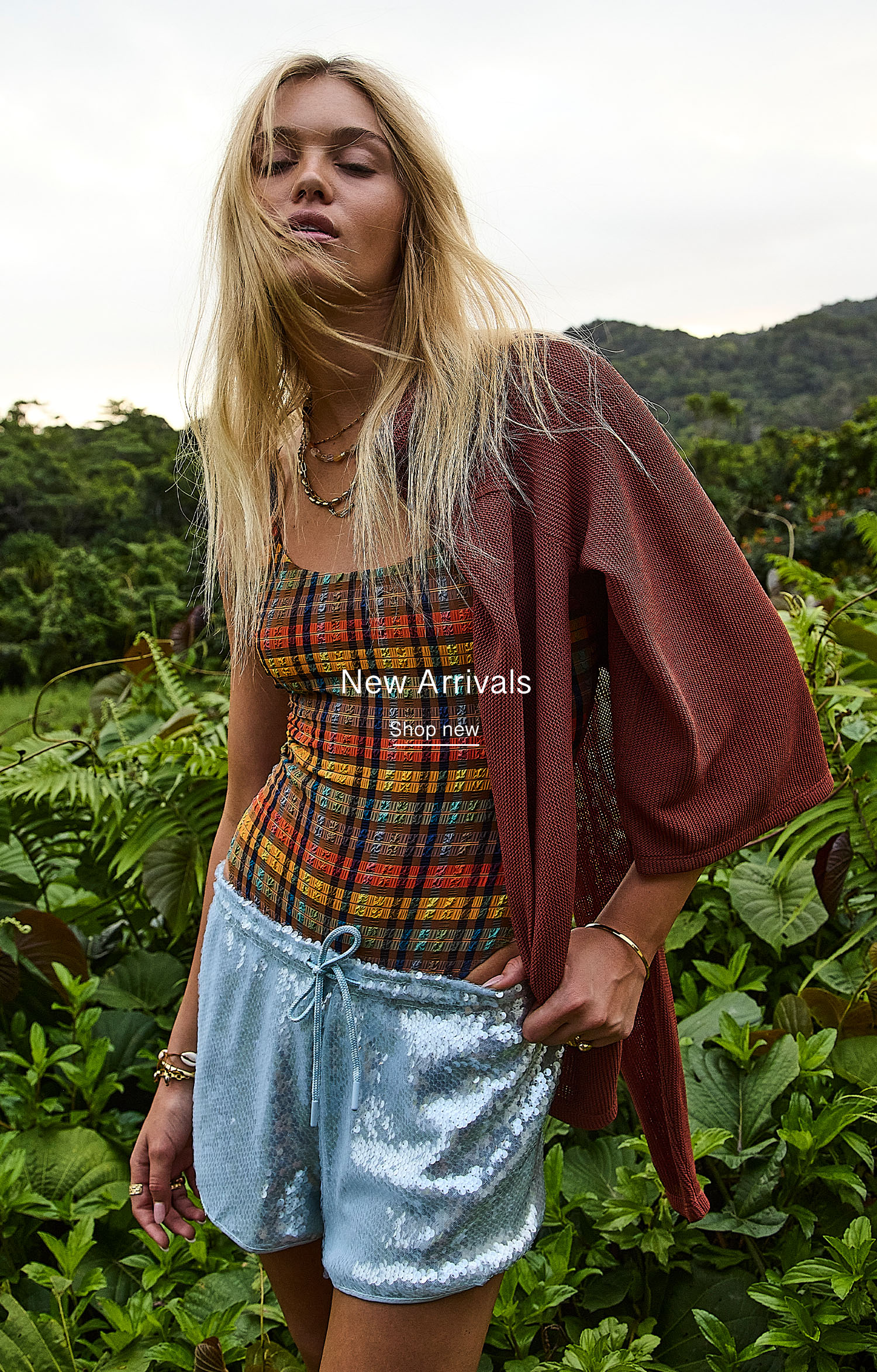 Model in a plaid top and shiny shorts among lush greenery.