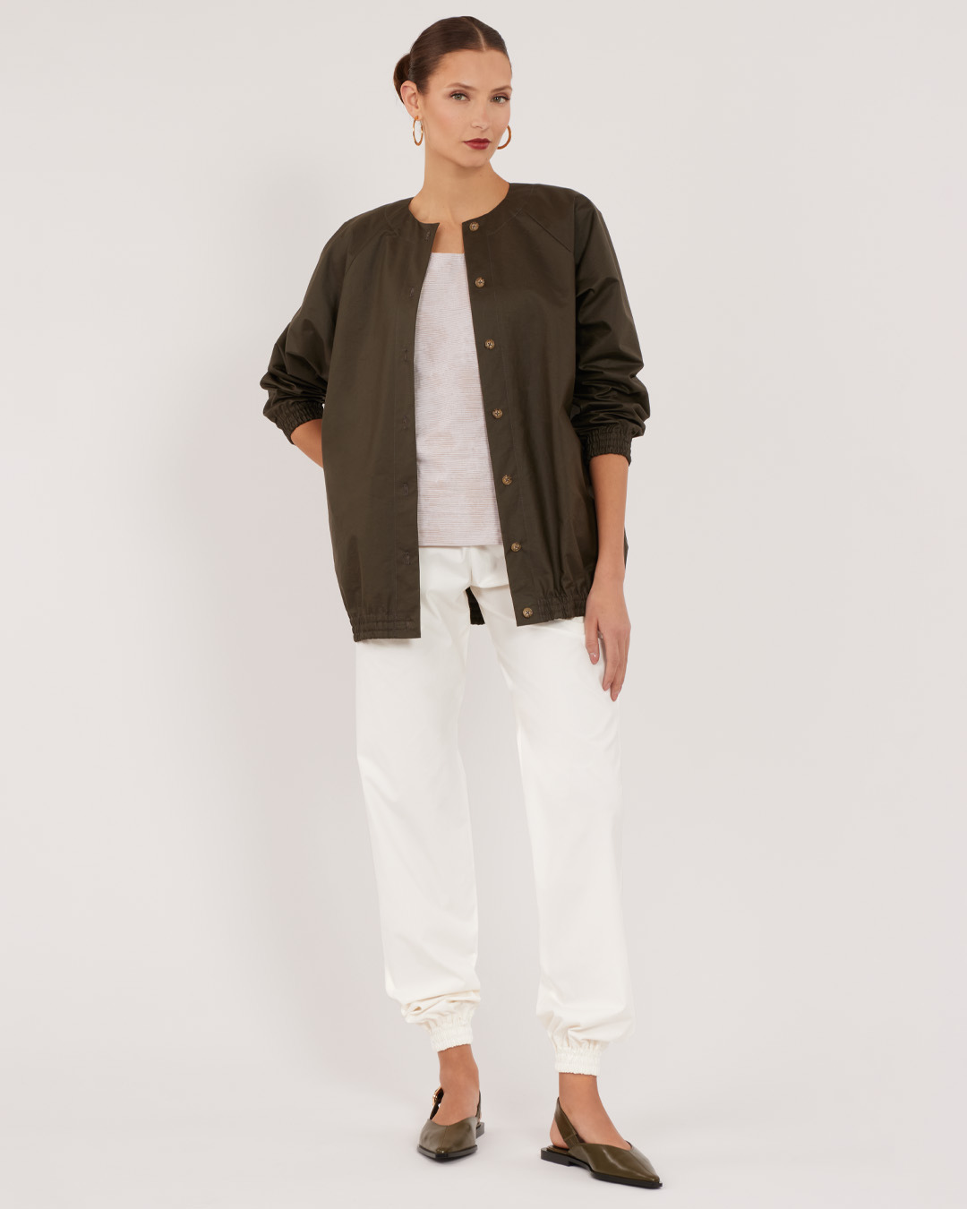 A woman wearing an olive jacket, white pants, and flats, standing against a plain background.