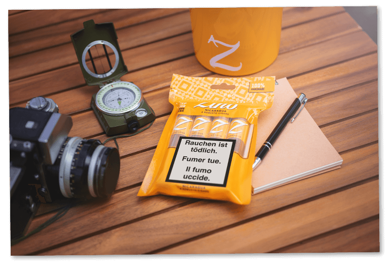 Vier Zino Nicaragua Robusto-Zigarren in ihrem Freshpack, platziert auf einem Holztisch neben einem Notizblock, einem Kugelschreiber, einer Zino Tasse und einem Kompass.