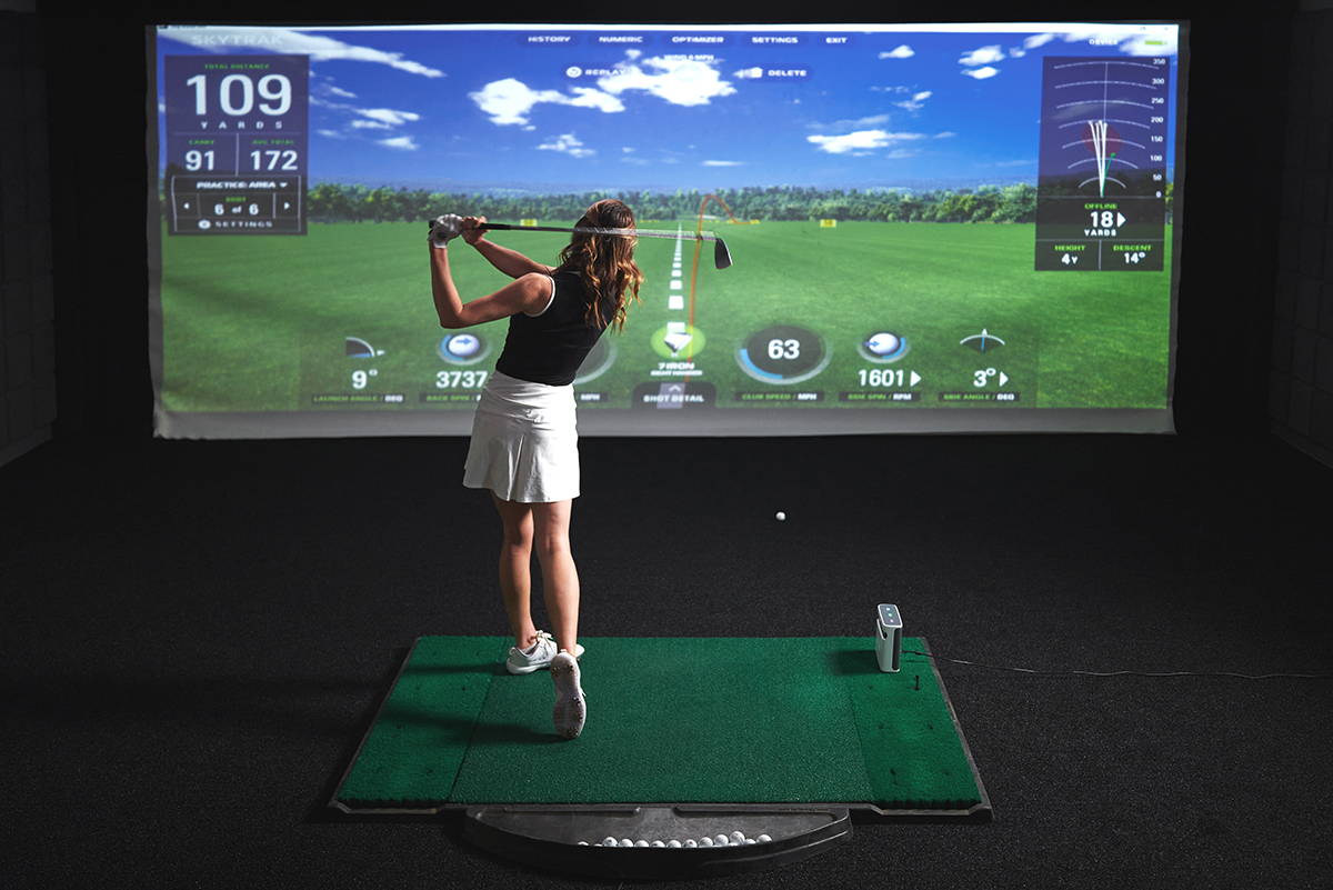A female golfer from behind just after swinging in a golf launch monitor with a SkyTrak+ and her shot data on the impact screen