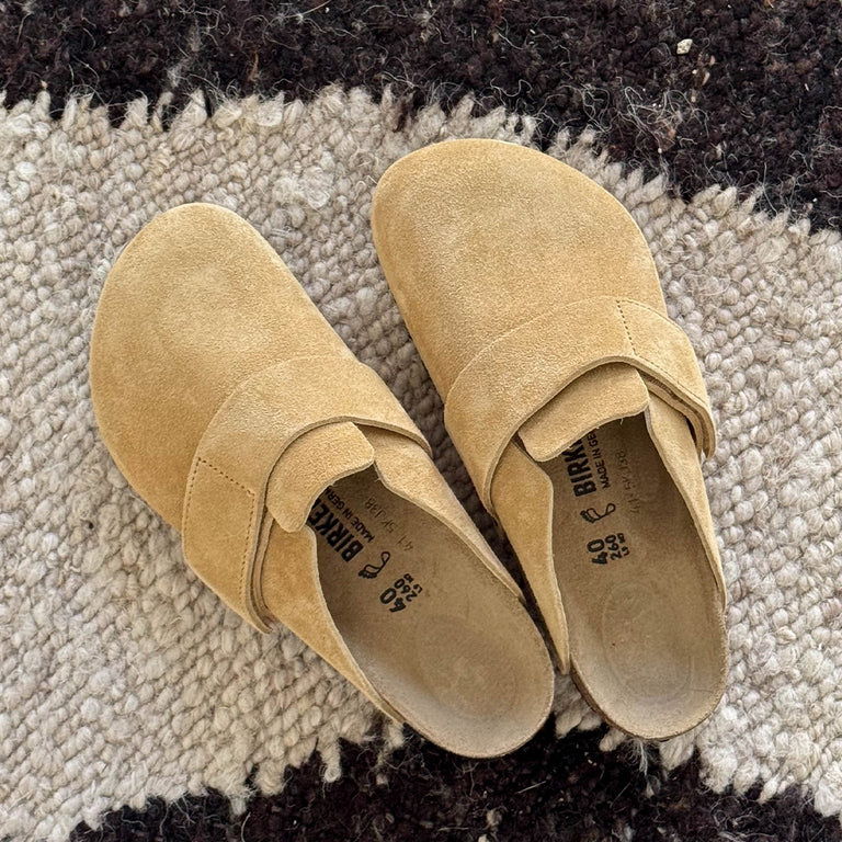 A pair of beige suede clogs on a textured rug.