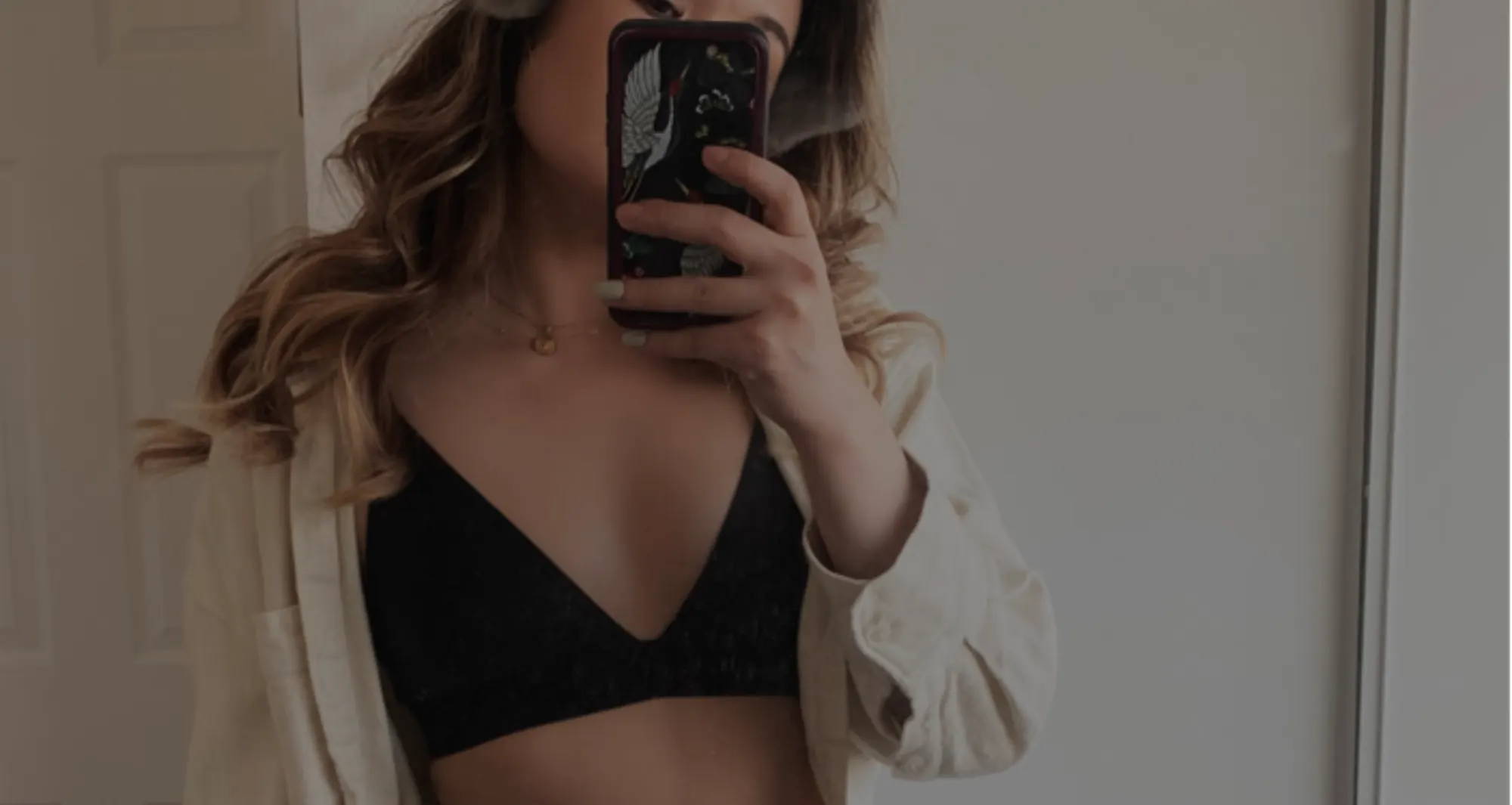  woman with long brown hair taking a mirror pic wearing a black triangle bralette, open cream shirt, and high rise jeans 