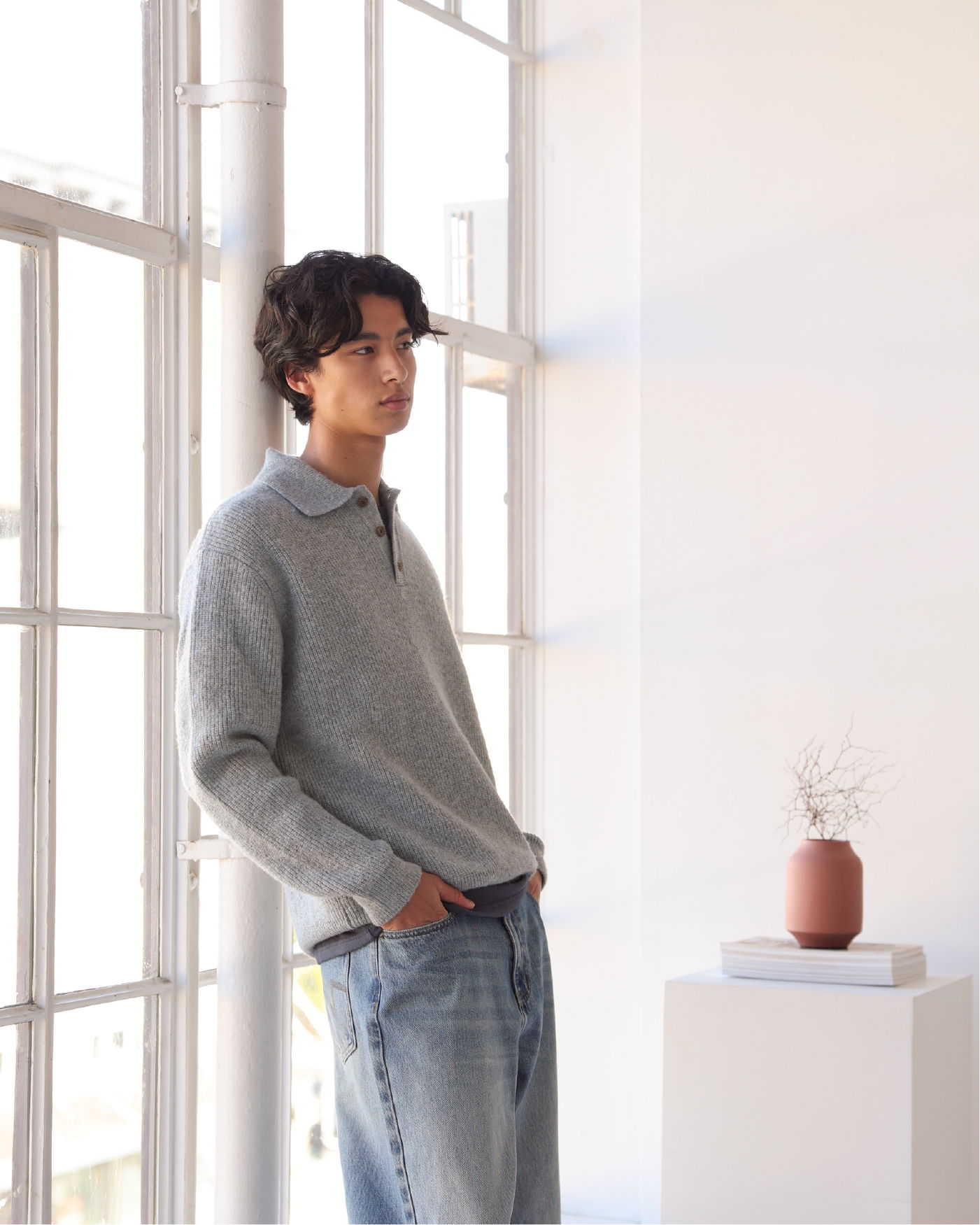 A young man in a gray sweater stands by a window.