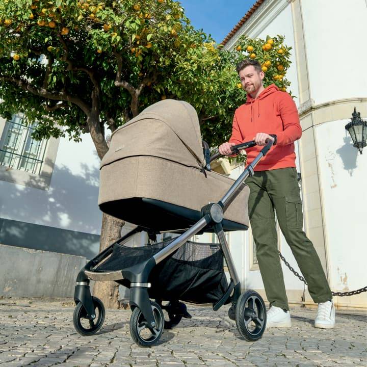 A dad wearing an orange jumper pushes his baby in a beige pushchair down a sunny cobbled street.
