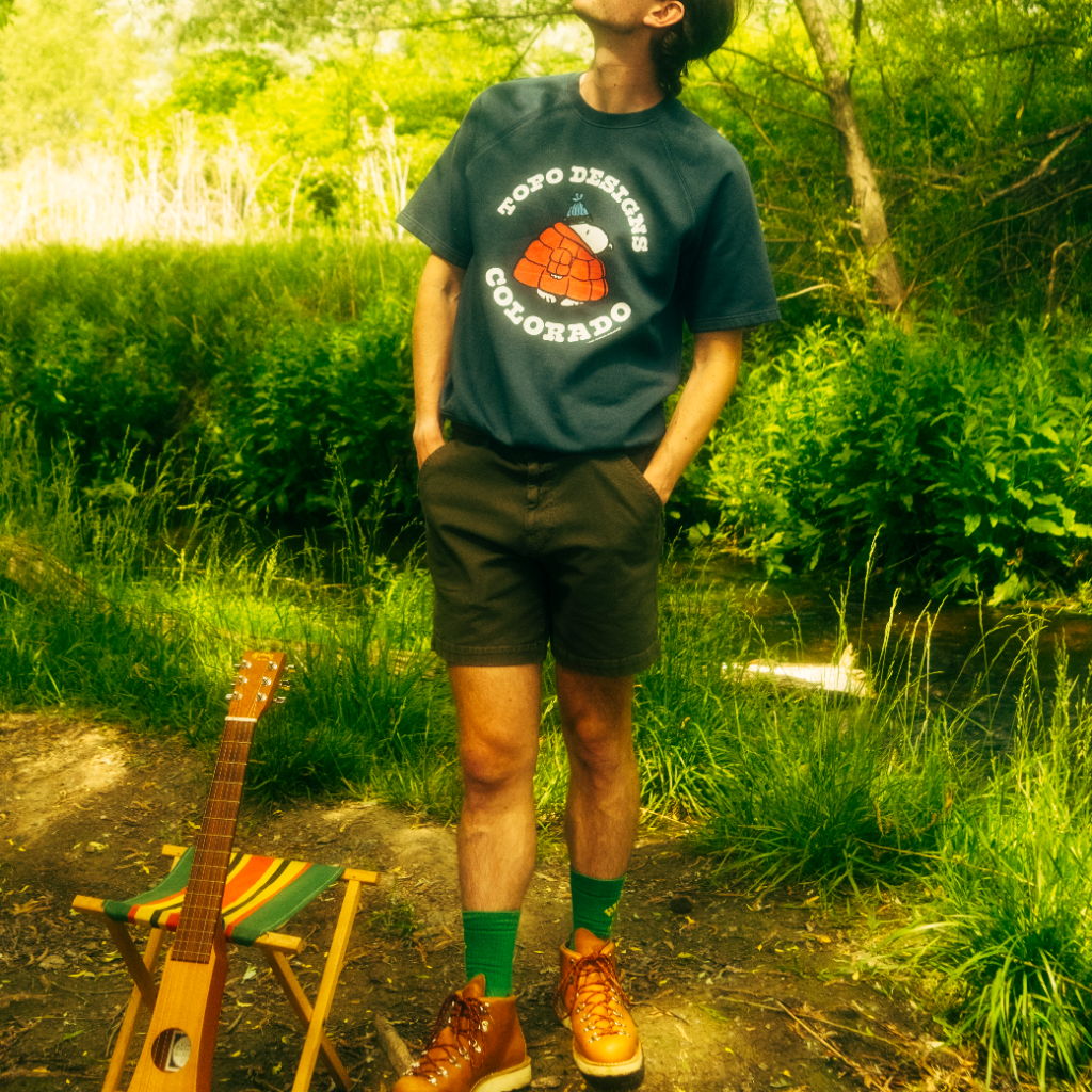 Person standing in nature, wearing a graphic t-shirt and shorts, with a guitar nearby.