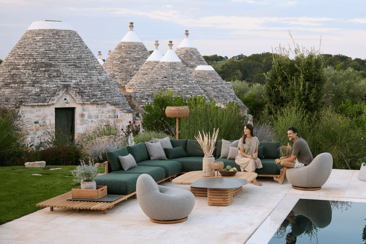 Outdoor living room with a deep green sectional and curved lounge chairs beside a pool, with stone cottages and lush landscaping behind.