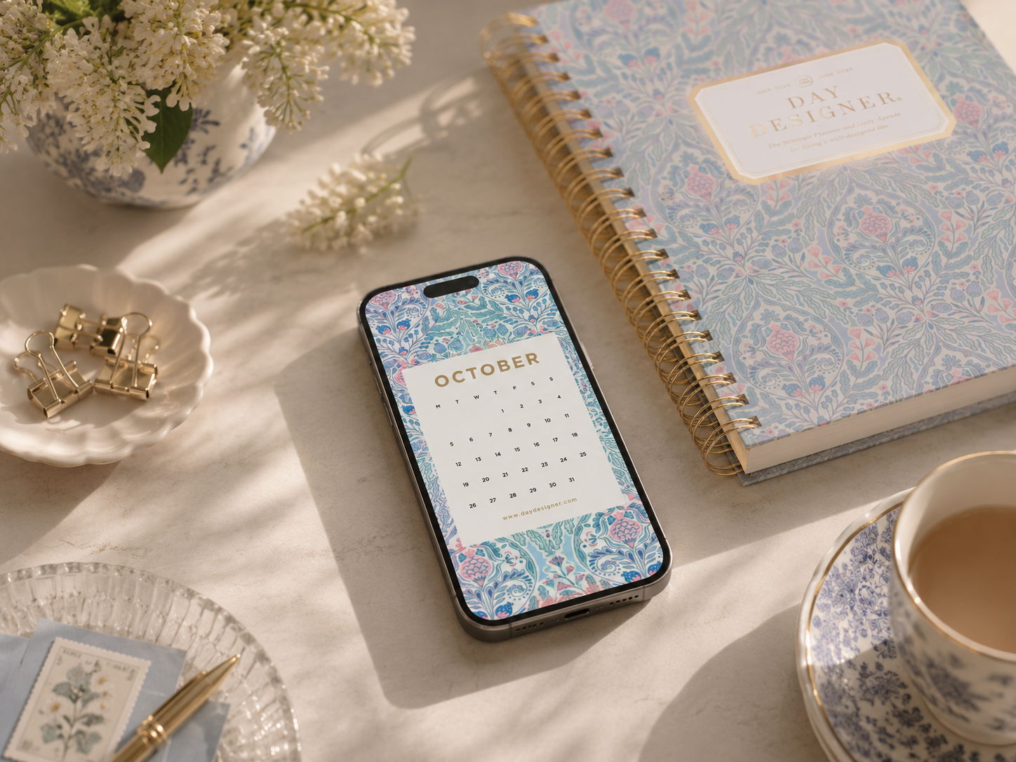 october 2026 digital wallpaper shown on mobile device, repeated blue and pink pattern, tea cup, day designer planner, dish with paper clips, vase with flowers