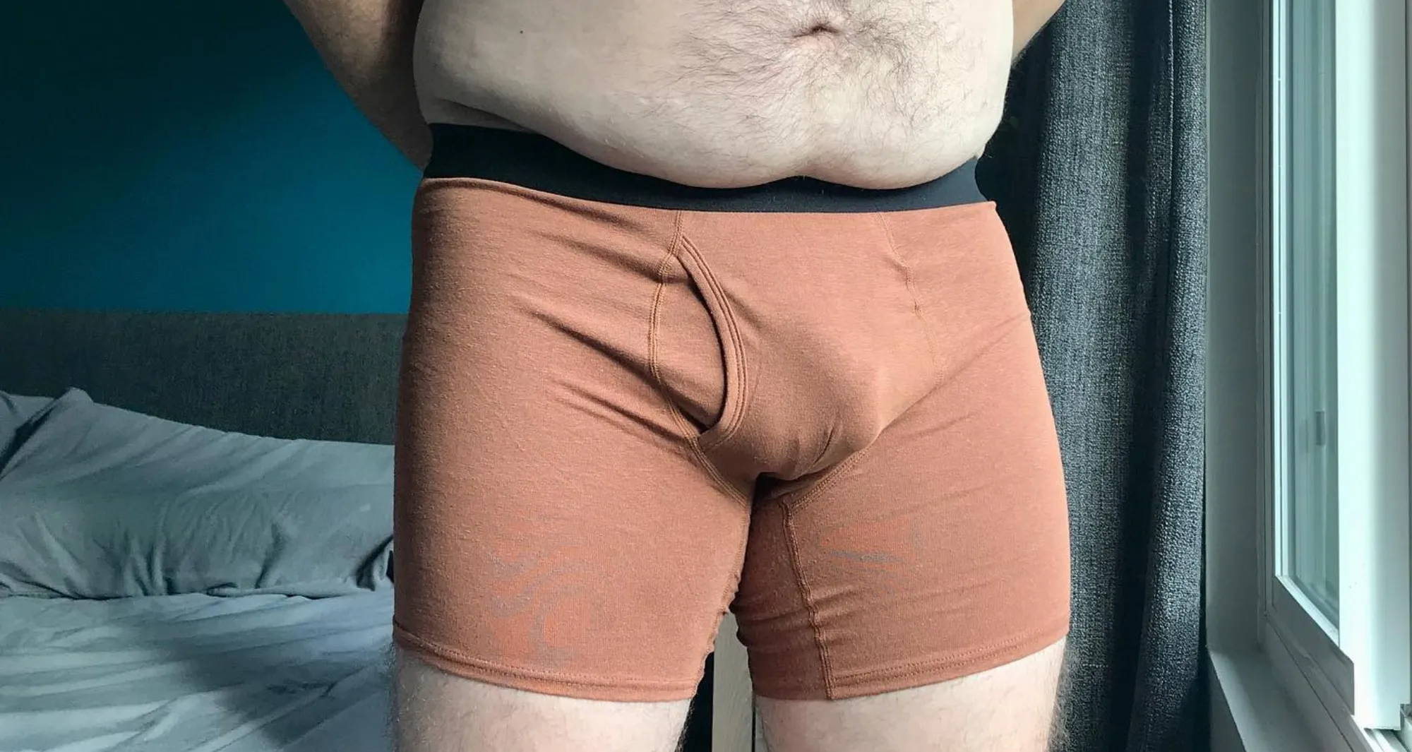 man’s lower half wearing a nude-orange pair of boxer briefs in a sunlit bedroom