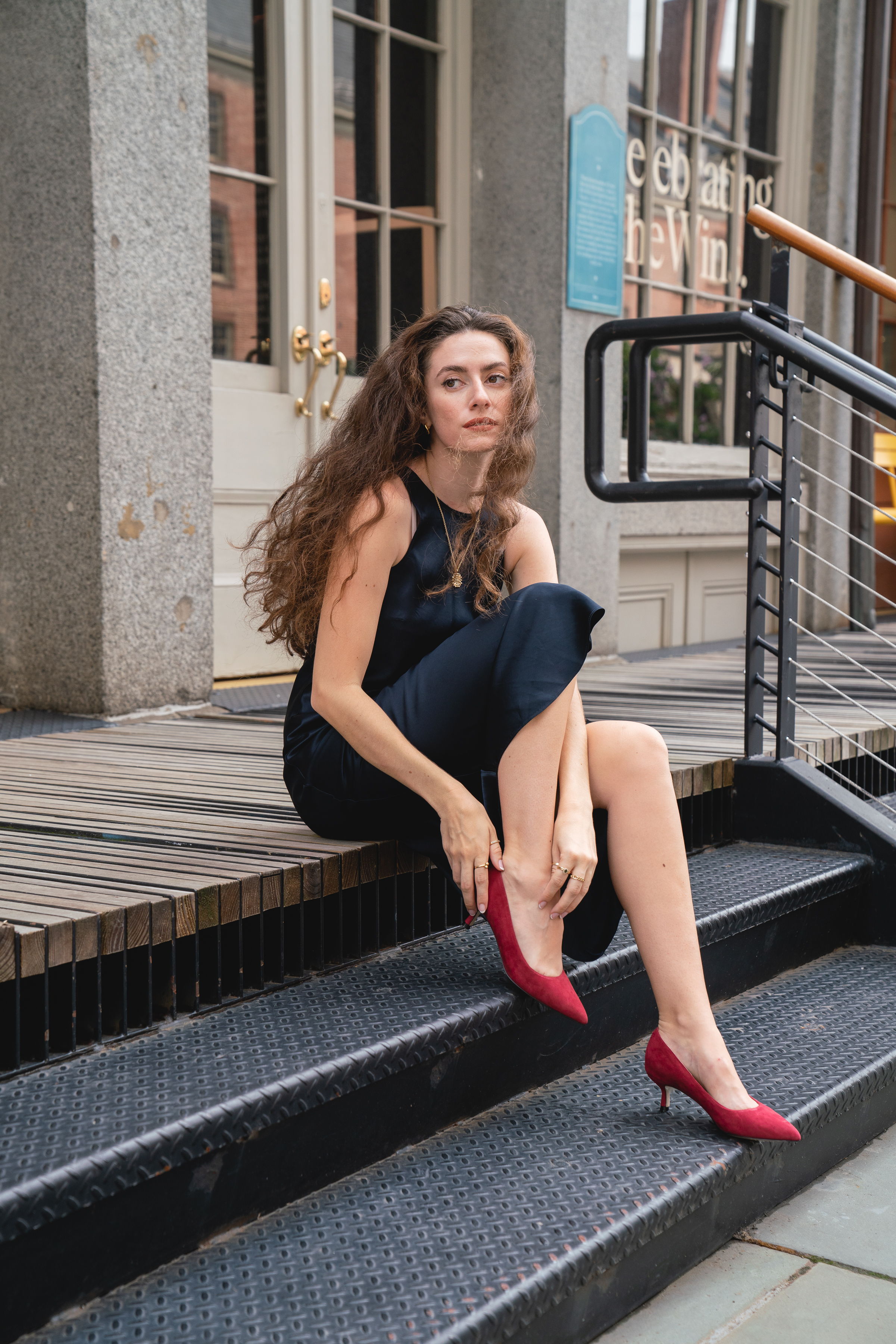 Woman sitting on stairs, adjusting her red high heels, with a casual outfit.