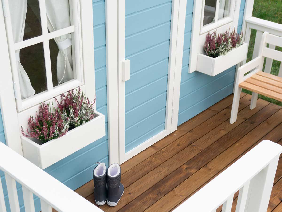Close up of white flower boxes and  wooden porch with rain boots of blue wooden playhouse by WholeWoodPlayhouses