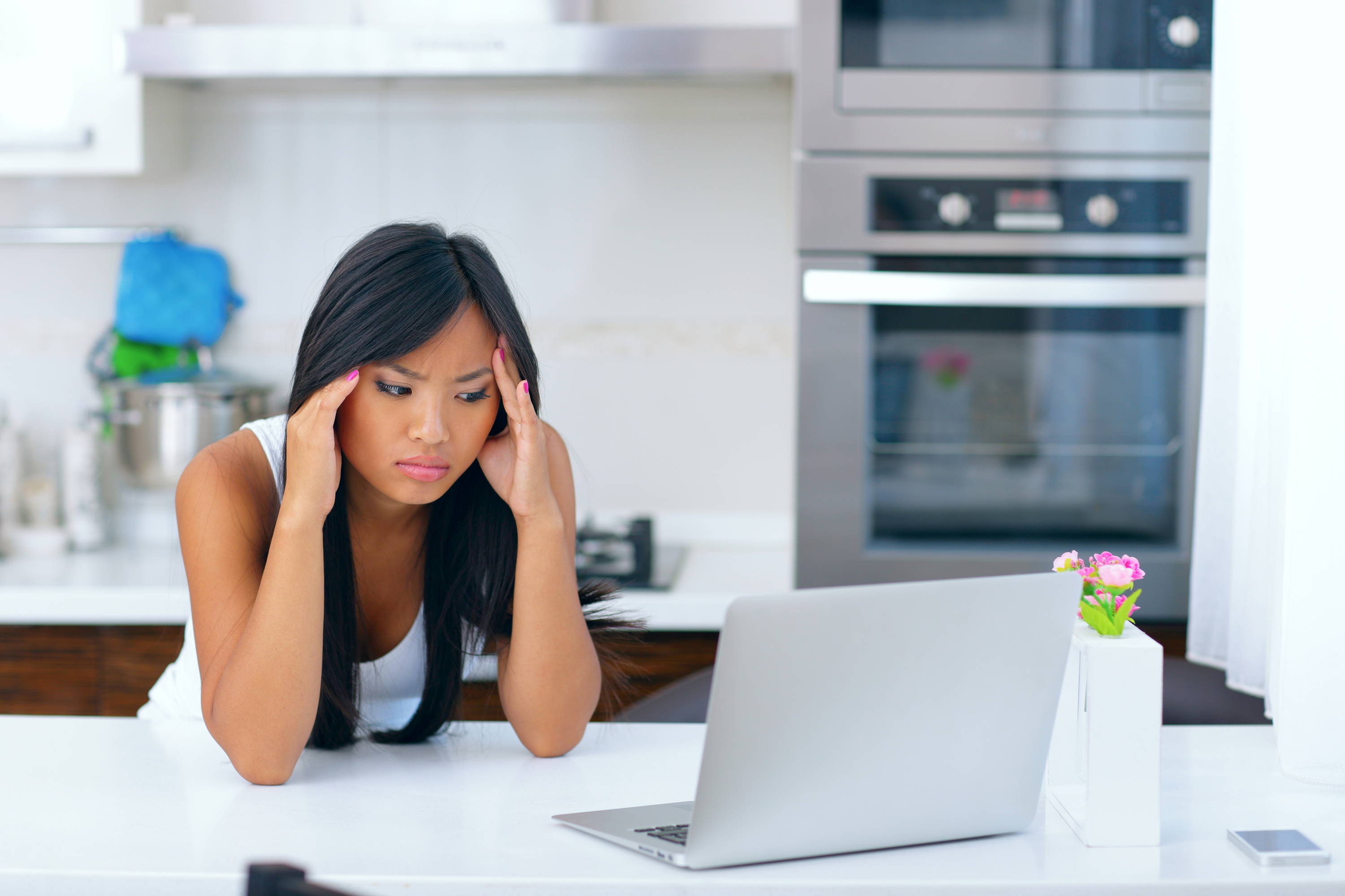 Woman holding her head with eyes open and elbows on a table. She is in the kitchen and staring at a laptop.