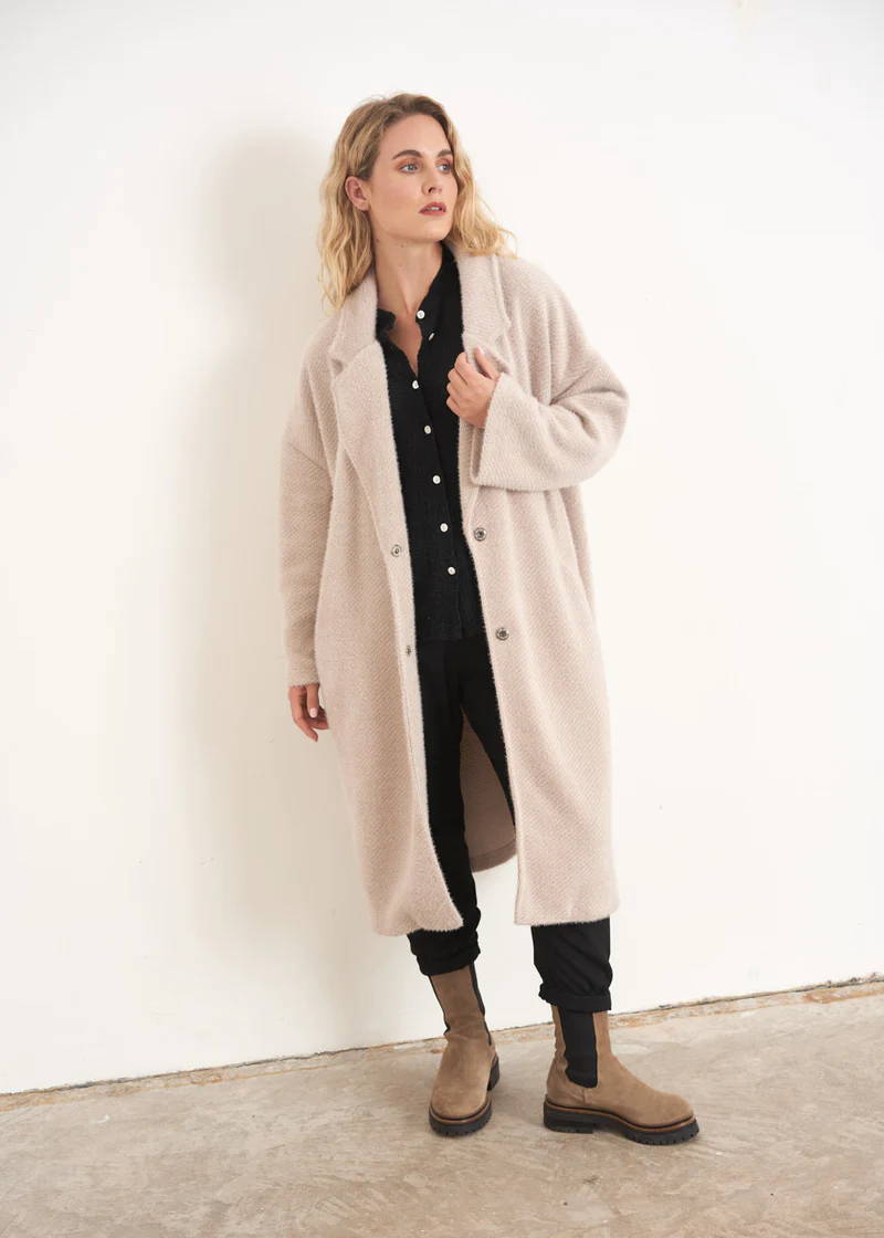 A model wearing a long, off white oversized winter coat over a black top, black trousers and taupe chelsea boots