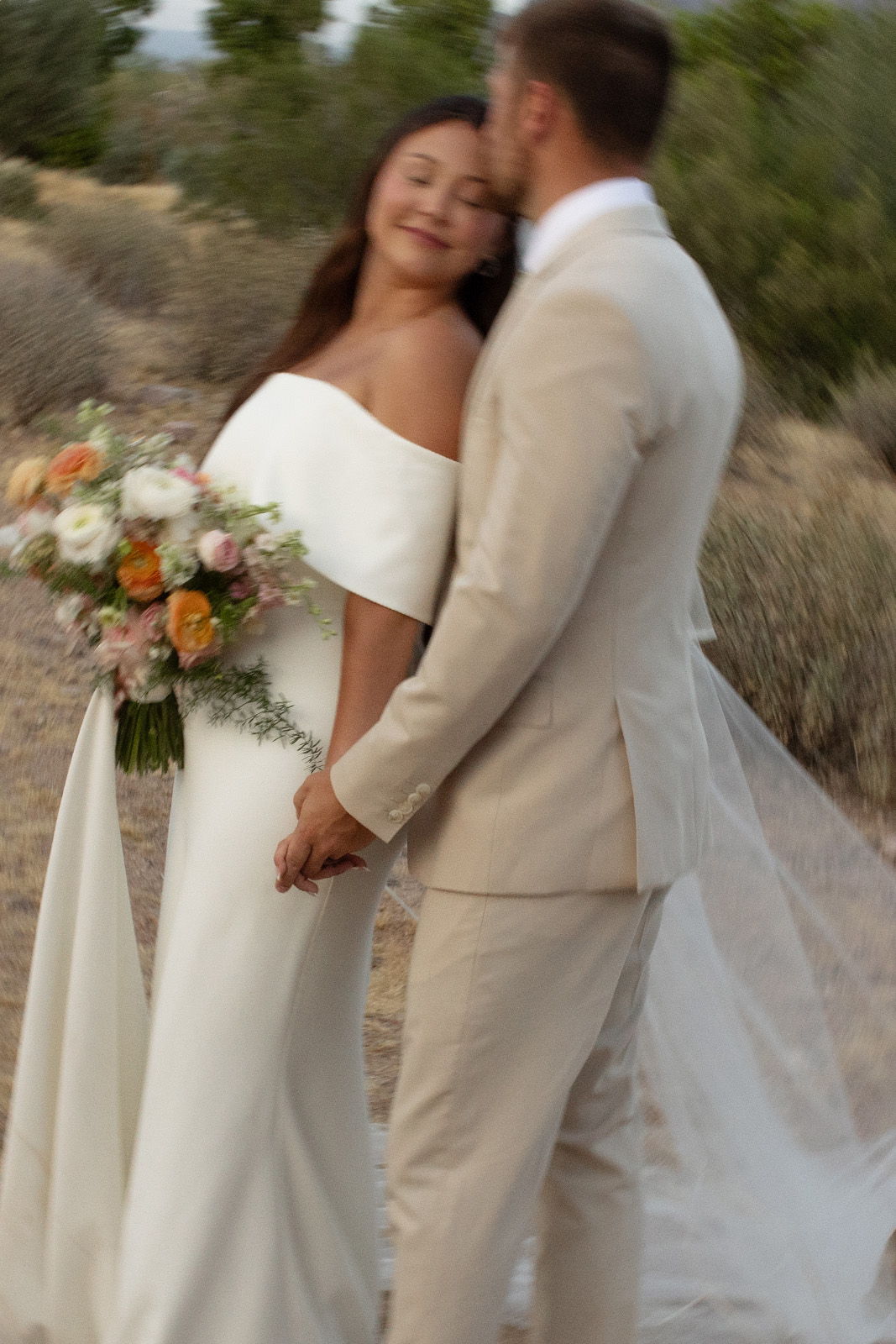 Bride and groom holding hands, smiling, surrounded by nature, wedding attire.
