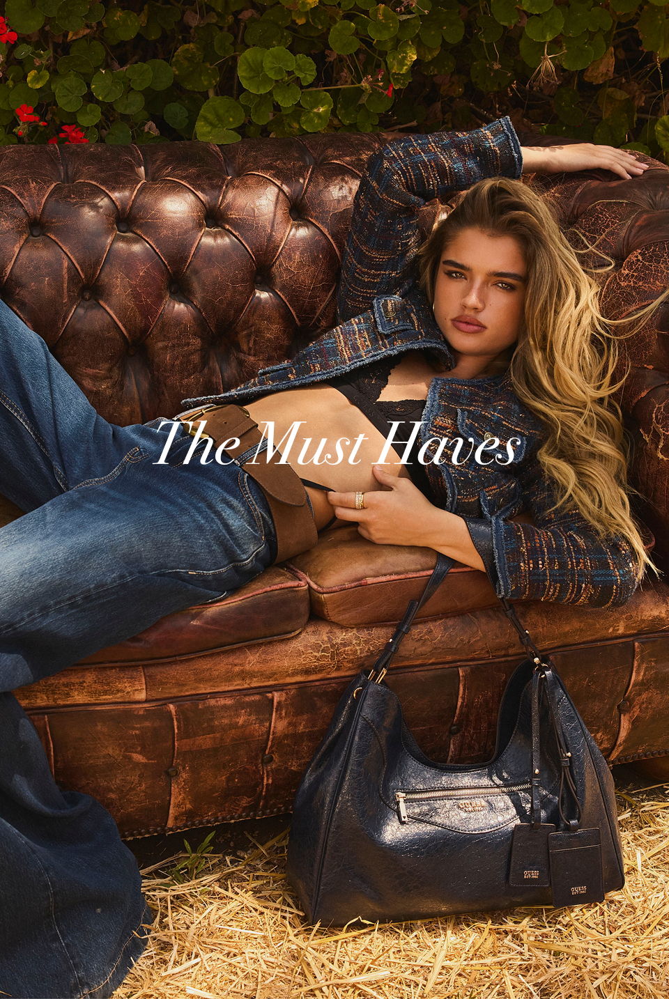 Woman lounging on a couch, wearing GUESS denim and holding a handbag.