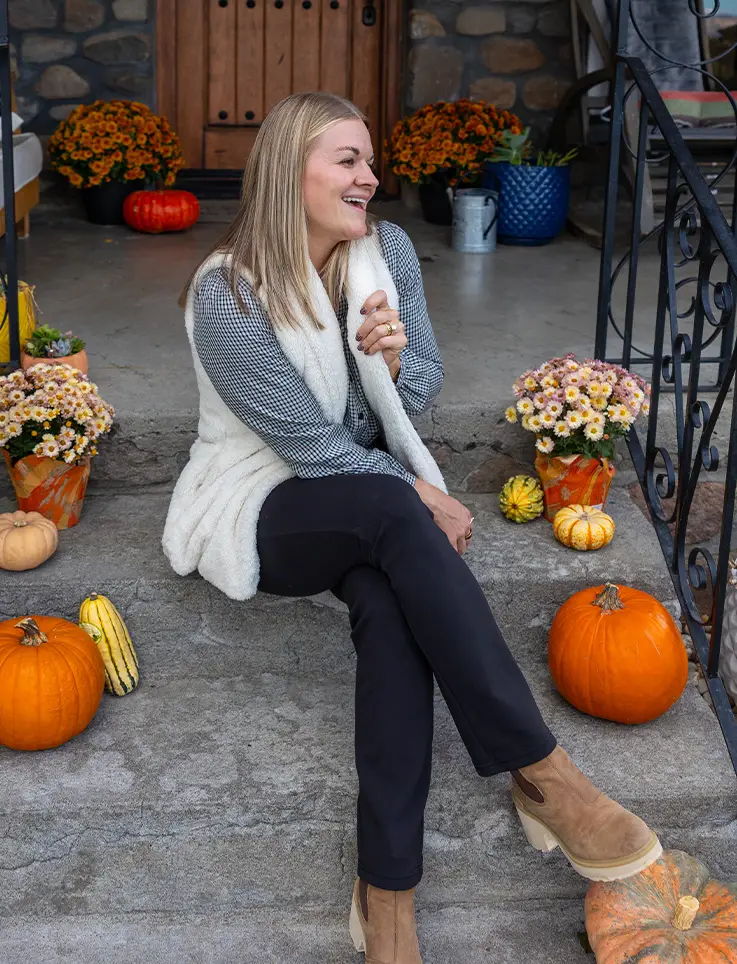 Smiling woman sitting on steps surrounded by pumpkins and flowers.