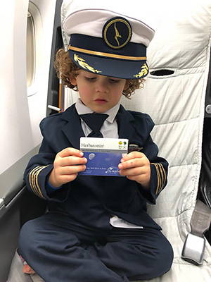 toddler sitting in airplane seat, wearing pilot uniform and holding box of herbatonin