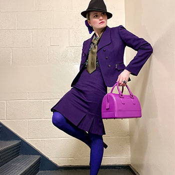 A woman dressed fashionably wearing a purple skirt suit and olive tie in an apartment stairwell