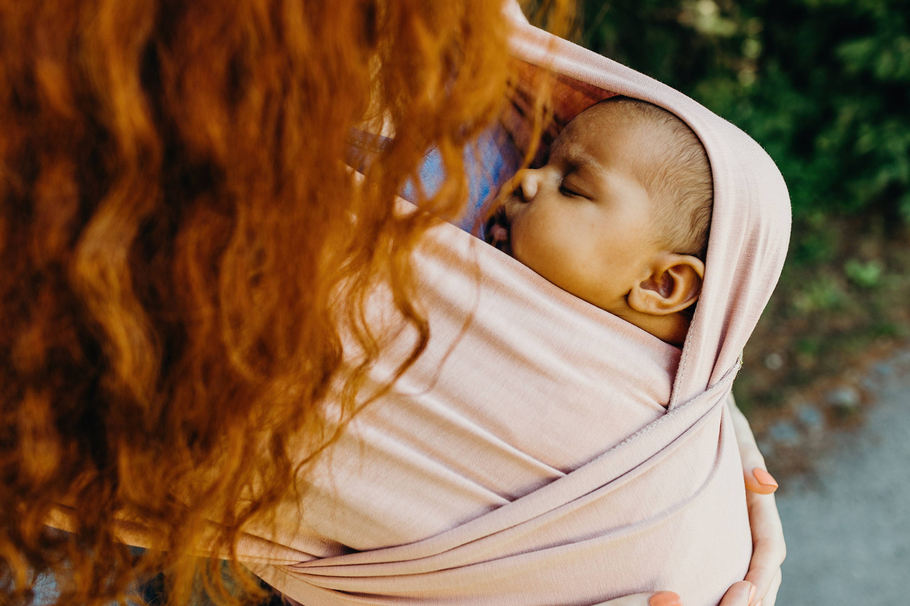 Small infant asleep in soft light pink bamboo baby wrap on red haired mother