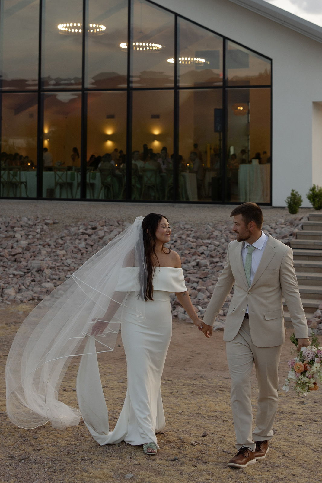 Bride and groom holding hands, walking outside a venue at sunset.