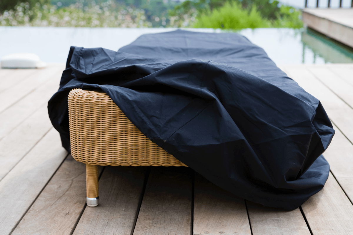 Woven outdoor lounger on a wood deck covered with a black protective furniture cover near a pool.