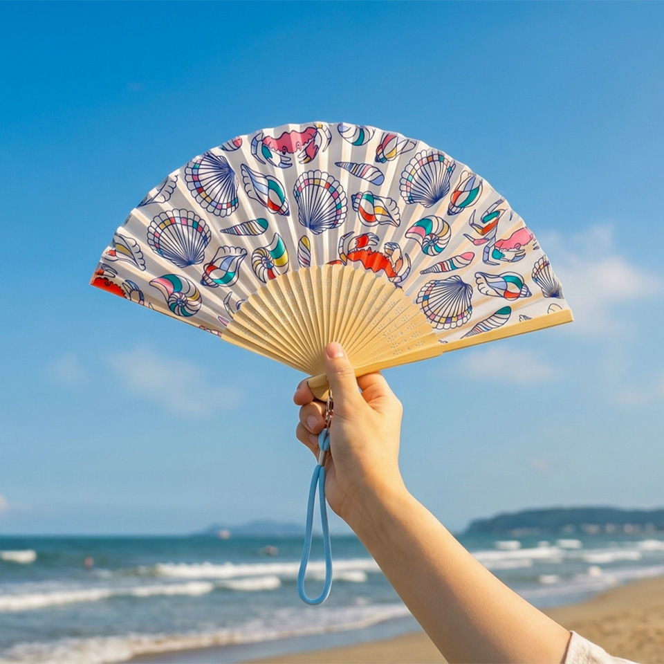 A hand holding a decorative fan against a beach and blue sky.