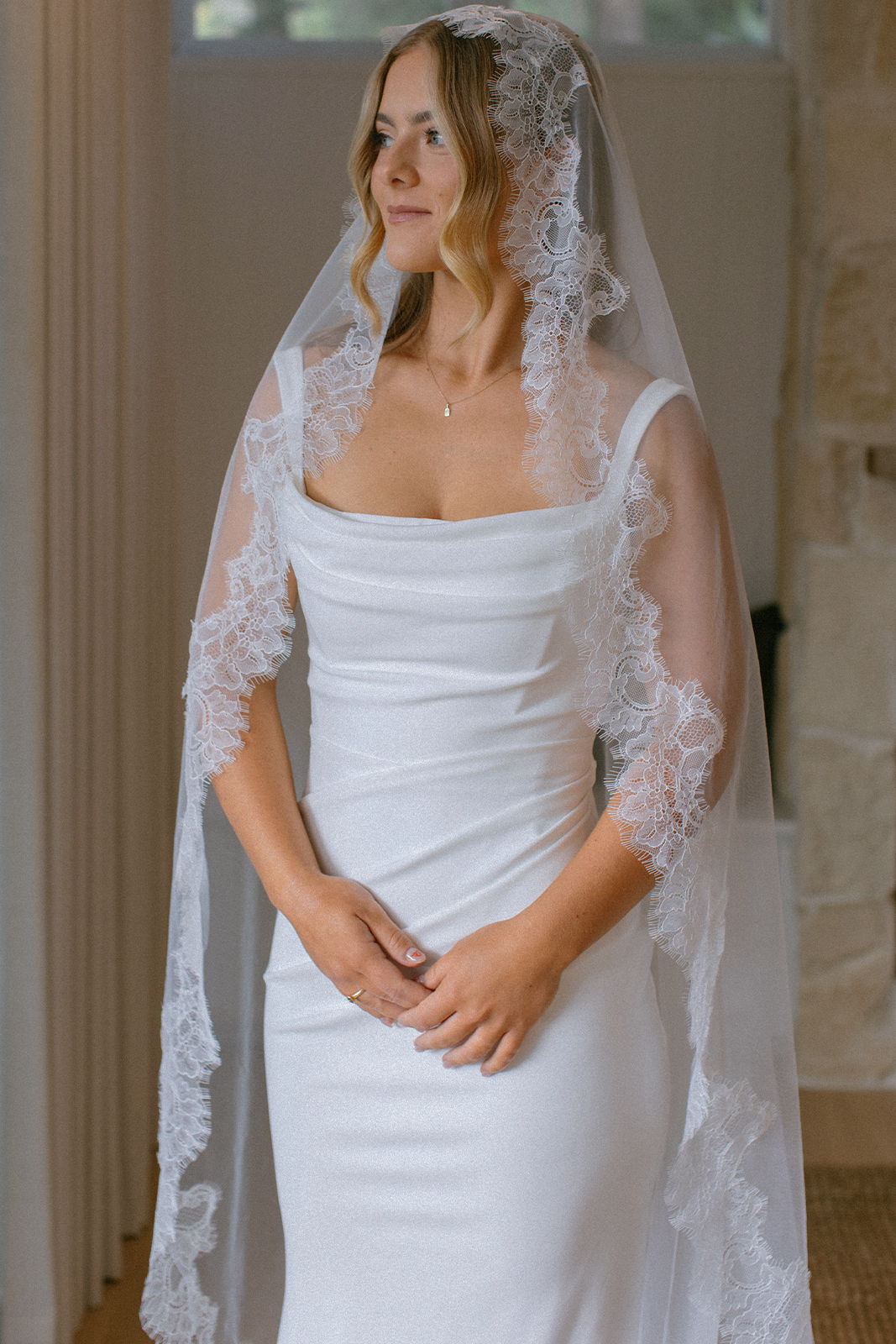 Bride in a white dress and lace veil, standing gracefully indoors.
