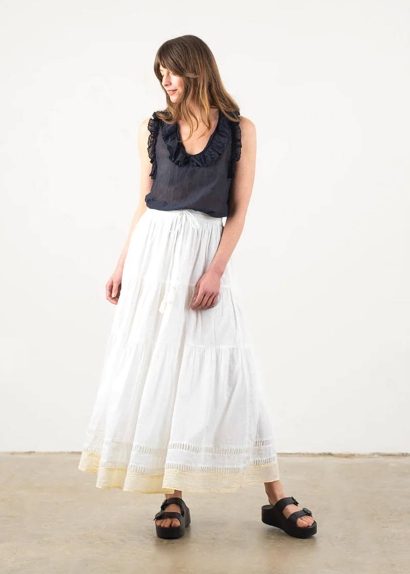 A model wearing a blue black sleeveless top with ruffle details around the neck and arms with a long, white maxi skirt and black platform slides.