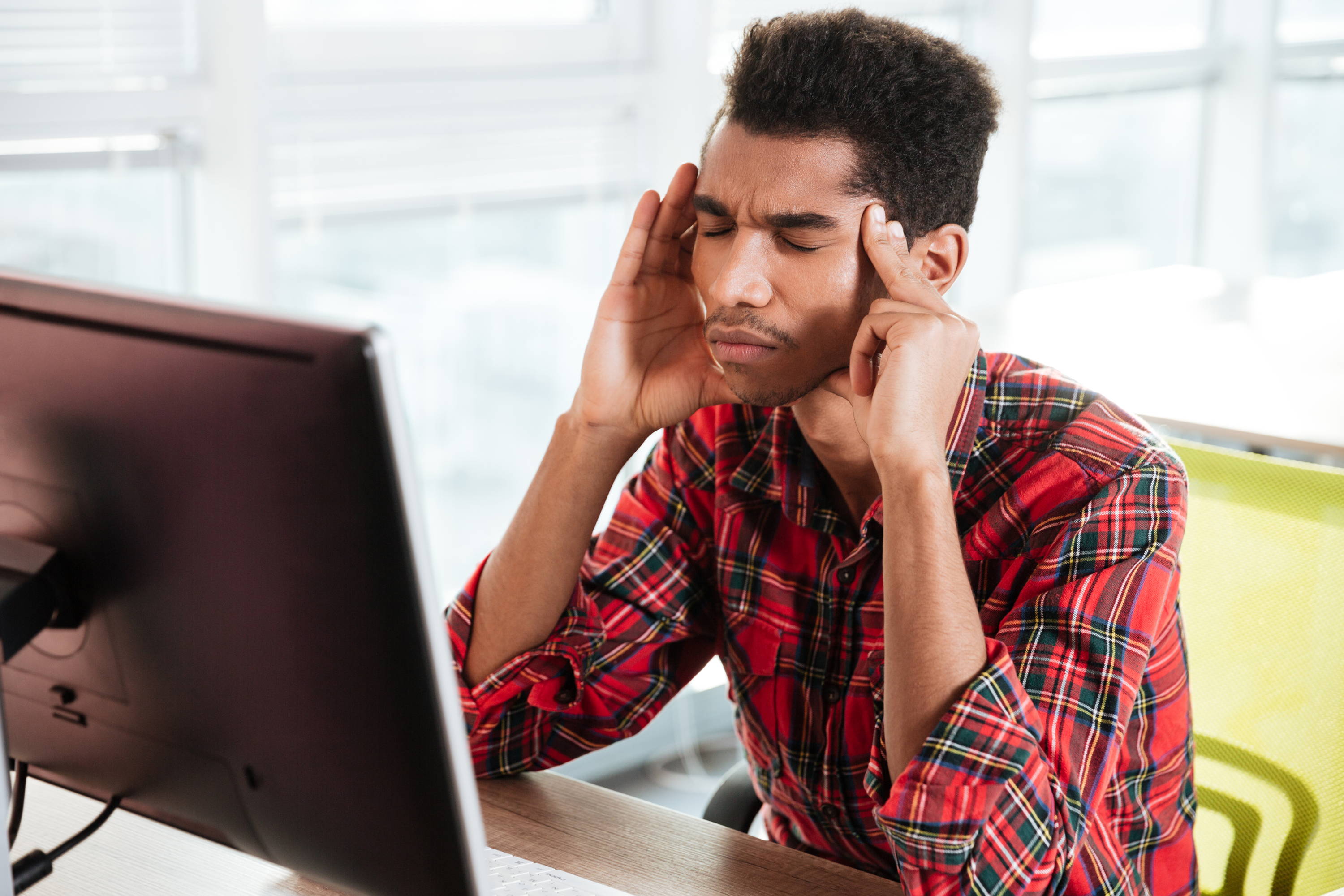 Man holing his temples wining with pain with his eyes closed in front of a computer.