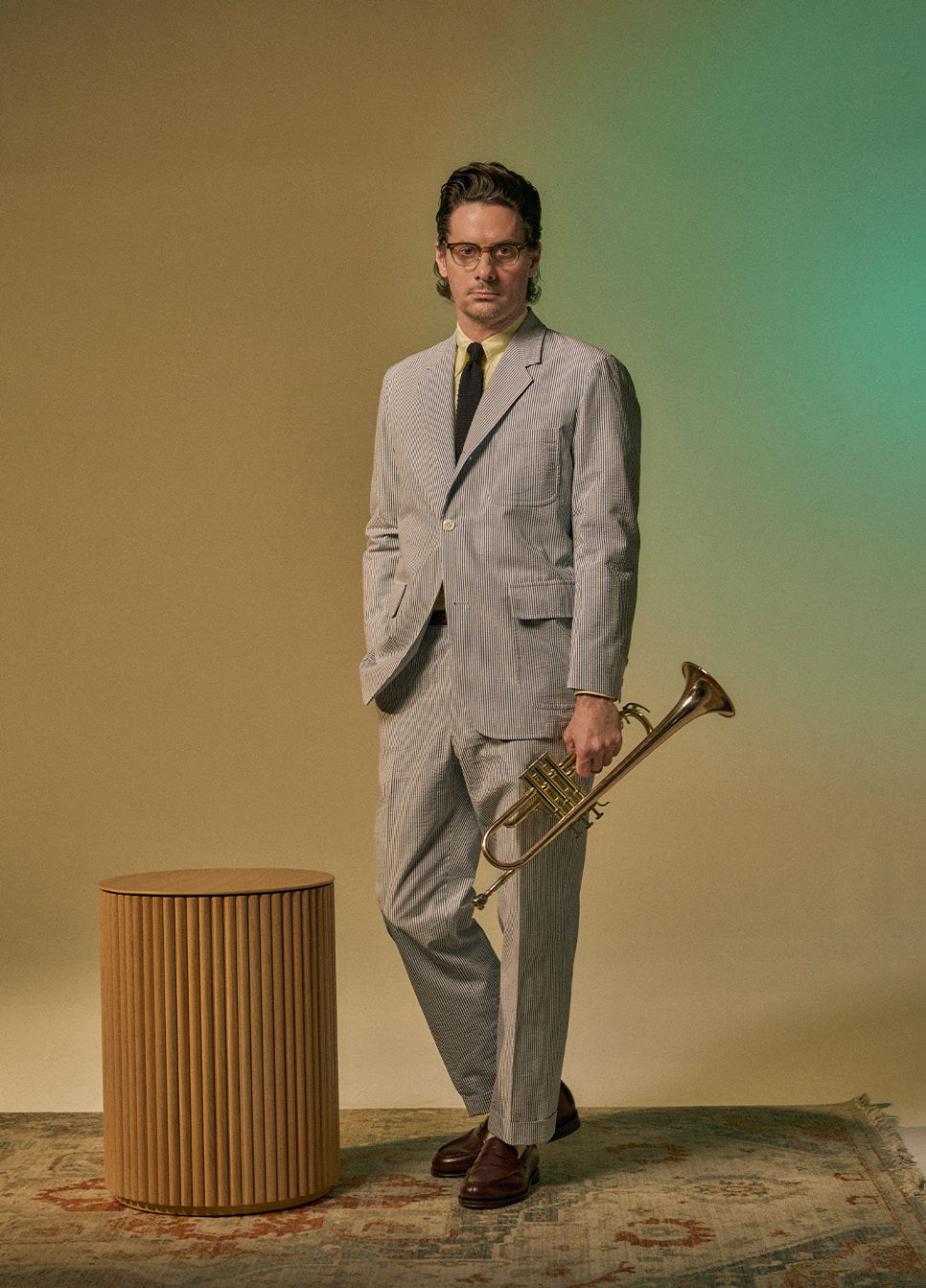 A man in a striped suit holding a trumpet, standing beside a wooden stool.