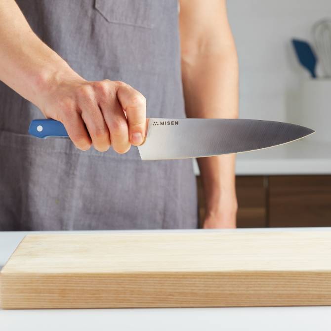 A chef holds a blue Misen Chef’s Knife towards the camera, with the blade’s steel and Misen logo clearly visible.