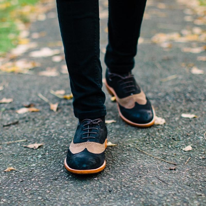 An outdoor lifestyle product shot from the calves down of a person walking forward on an asphalt path scattered with autumn leaves. The person is wearing slim-fit black jeans and distinctive two-tone leather wingtip oxford shoes featuring dark navy or black panels on the toe and heel, contrasting with a light tan or beige perforated saddle overlay.