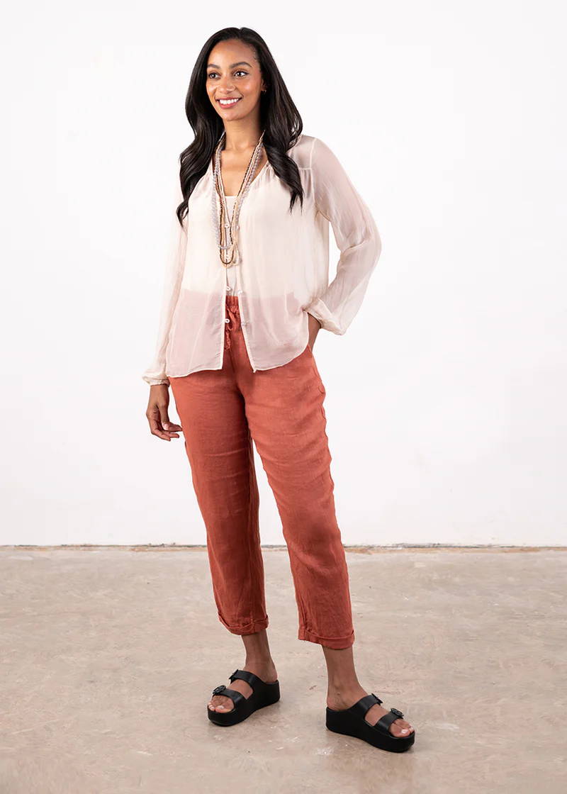 A model wearing a semi sheer, light pink shirt over a white top, terracotta coloured linen trousers, black chunky platform slides and a collection of beaded crystal necklaces