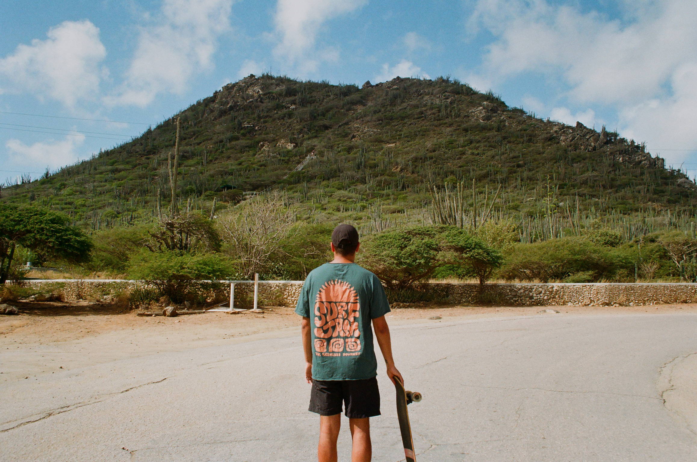 A man looking up a mountain in Aruba holding his skateboard