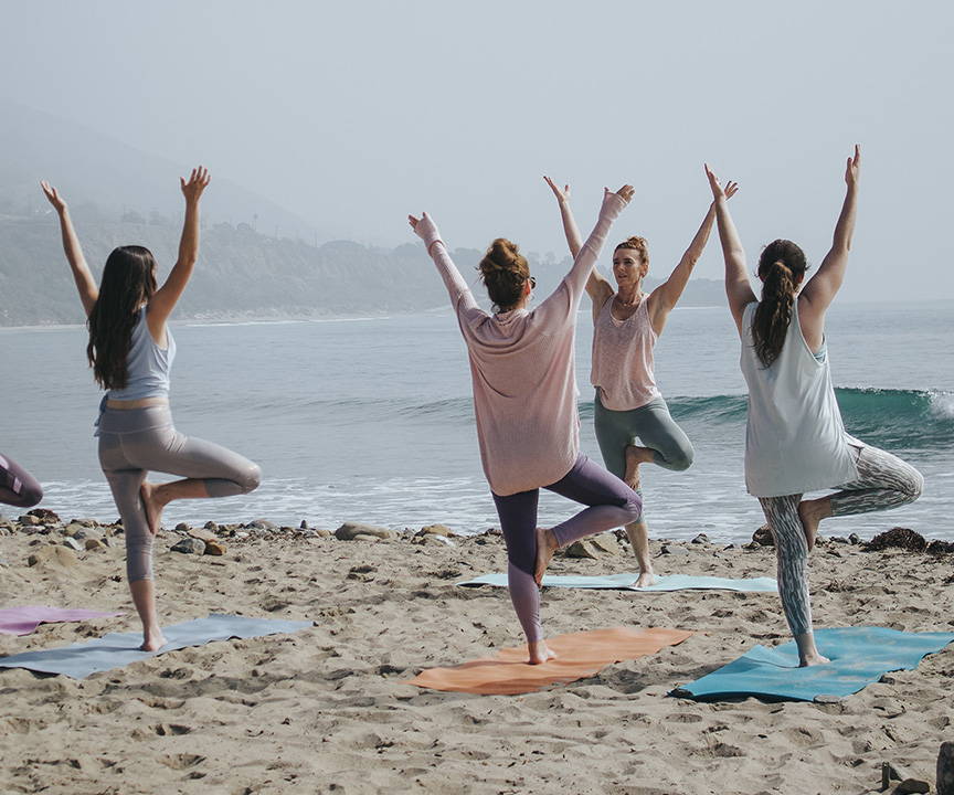 A group of three women and their yoga instructor doing yoga in the sand on a beach.