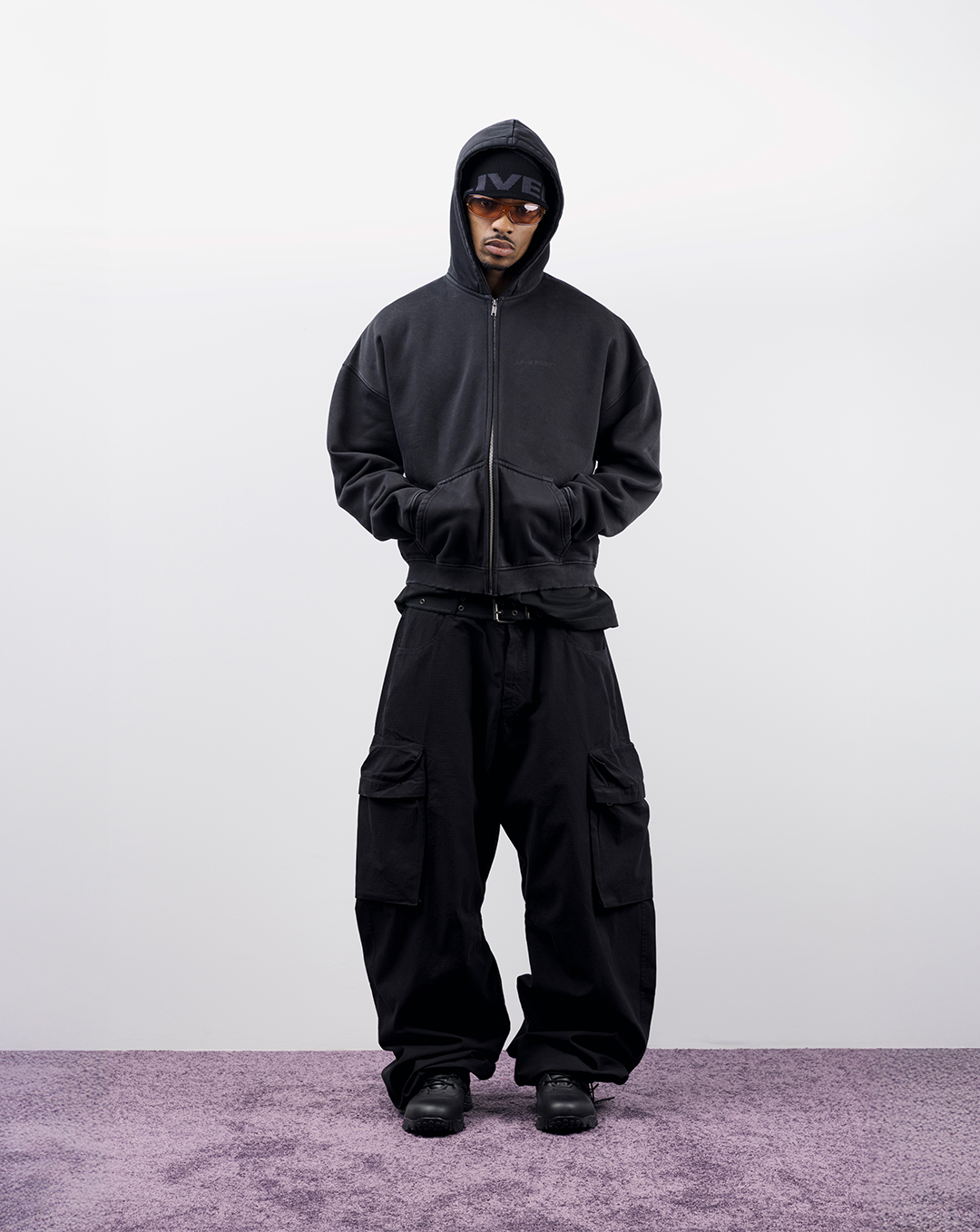 Person wearing black hoodie and pants, standing on purple carpet against white background.