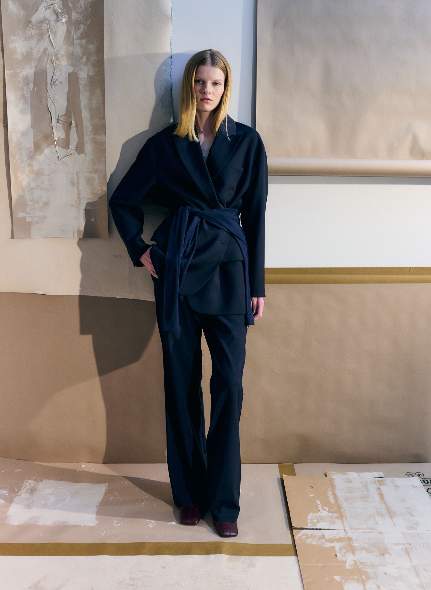 Model in a black outfit stands against a backdrop of craft paper.