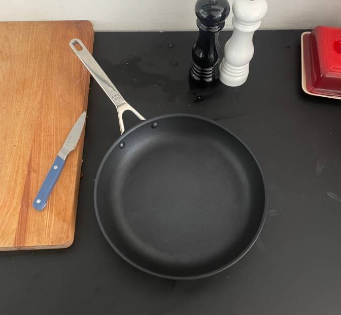A bird’s eye view of a Misen Nonstick Pan on a black countertop. To the left of the pan is a blue Misen Paring Knife.An unseen hand uses a wooden spoon to stir chopped red onion in a Misen Nonstick Pan, which sits on a stovetop.