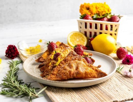 Fried pastries on a plate with fruits and flowers in the background.